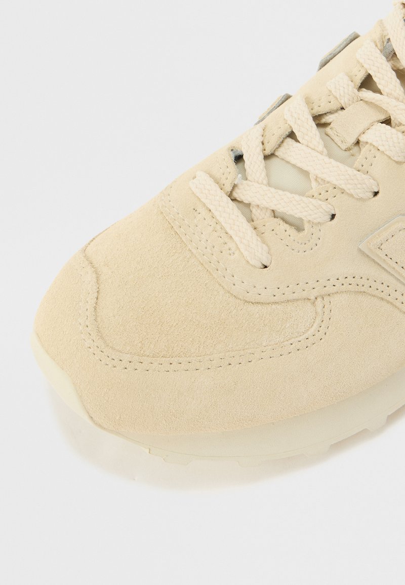 Zapatilla de ante beige con parte superior texturizada, punta redondeada y cordones planos. Presenta una suela de color claro y detalles de costura sutiles.