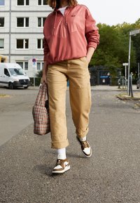 Světle růžová mikina s kapucí a půlzipem, béžové cargo kalhoty a hnědé-bílé tenisky. Nese kostkovanou tašku.