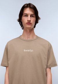 Light brown cotton T-shirt with short sleeves and a round neckline. Features white branded logo on the chest and a smooth texture.