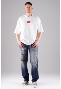 Weißes übergroßes T-Shirt mit rotem Logo, kombiniert mit distressed blauen Jeans und grünen Canvas-Sneakers, vor einem neutralen Hintergrund.