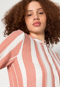 Camisa de malha com padrões verticais em laranja e branco, tecido texturizado, fechamento por botão e gola.