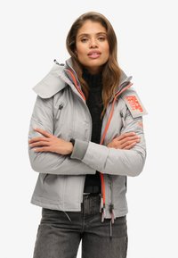 Veste isolée grise avec des accents orange, col haut et multiples zippers. Poignets côtelés et poches latérales sont présents.