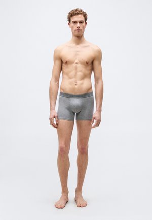 Young man with curly hair standing barefoot, wearing gray boxer briefs with a branded waistband, against a plain light background.