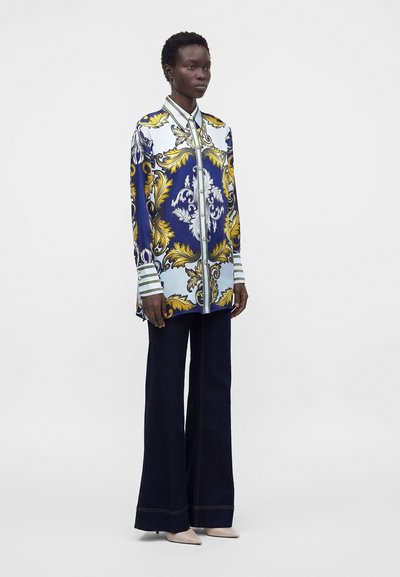 Femme portant une longue chemise à motifs bleue, blanche et dorée, un pantalon large foncé et des chaussures beige à bouts pointus, debout devant un fond uni.