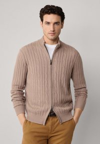 Beżowy sweter zapinany na zamek z dzianiny w warkocze, z wysokim kołnierzem i prążkowanymi mankietami, z wyróżniającymi się wzorami i gładką teksturą. Noszony z beżowymi spodniami.