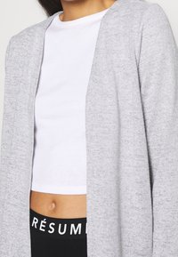 Cardigan aperto grigio realizzato in morbido tessuto a maglia, con una vestibilità comoda. Indossato sopra una maglietta bianca a maniche corte e pantaloni neri con scritte bianche.