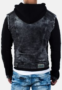 Felpa nera abbinata a una giacca di jeans grigia sbiadita. Presenta bordi sfilacciati, maniche di colore contrastante e un'etichetta con logo sul fondo.