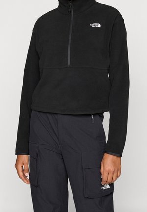 Persoon draagt een zwarte North Face fleece met een kwart ritssluiting en een zwarte cargobroek met voorvakken met klep, beide met merklogo's.