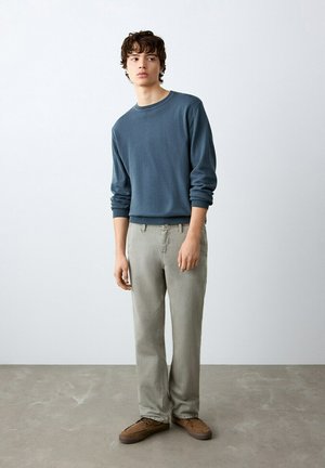 Mladić stoji u zatvorenom prostoru noseći plavu košulju dugih rukava, svijetlosive hlače i smeđe cipele od brušene kože na betonskom podu.