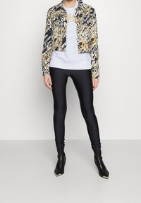 Giacca cropped nera e oro a fantasia sopra un maglione bianco, abbinata a leggins neri e stivaletti alla caviglia neri con accenti dorati.