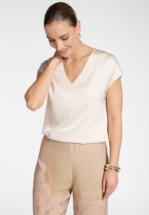 Donna con capelli pettinati all'indietro, che indossa una camicetta a maniche corte di colore beige chiaro e pantaloni beige con un delicato motivo floreale, accessoriata con braccialetti.