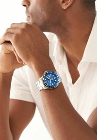 Montre en acier inoxydable avec un cadran bleu et une lunette noire, présentant des accents argentés et des index en bâton pour les heures. Portée à un poignet, partiellement visible.