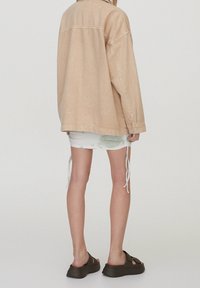 Veste en denim beige à coupe décontractée, associée à une mini-jupe blanche tie-dye avec des détails à cordon, et des sandales compensées marron.