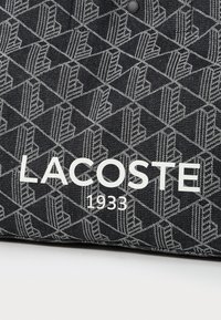 Țesătură neagră cu un model geometric, evidențiată de accente albe. Logo-ul "LACOSTE 1933" este afișat proeminent în text alb.