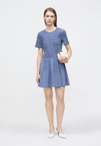 Robe courte à manches courtes à carreaux bleus avec un corsage smocké et une jupe évasée, associée à une pochette blanche et des talons pointus blancs.