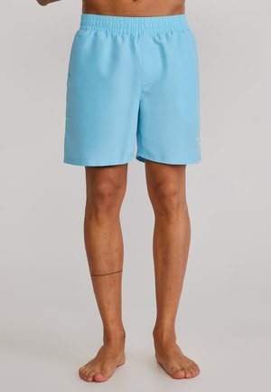 Moški v svetlo modrih kopalkah stoji bosi pred enobarvnim ozadjem.