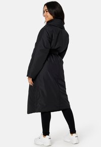 Zwarte lange pufferjas met een riem in de taille, hoge kraag en opgestikte zakken. Het materiaal heeft een gladde textuur met subtiele stiksels.