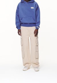 Sudadera azul con capucha y bolsillo tipo canguro; pantalones cargo beige con múltiples bolsillos laterales; zapatillas crema. Conjunto simple y casual.