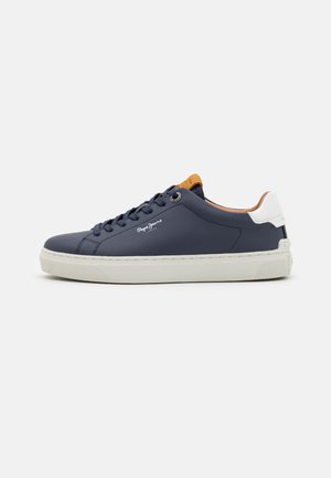 Zapatilla de deporte azul marino con detalles blancos, diseño de cordones, textura suave, suela de goma y detalle del logo en el costado. Parte superior de cuero suave.