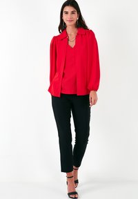 Red blouse with puffed sleeves and a collar, paired with black pants and strappy sandals. Skin is shown, hair is wavy and dark.