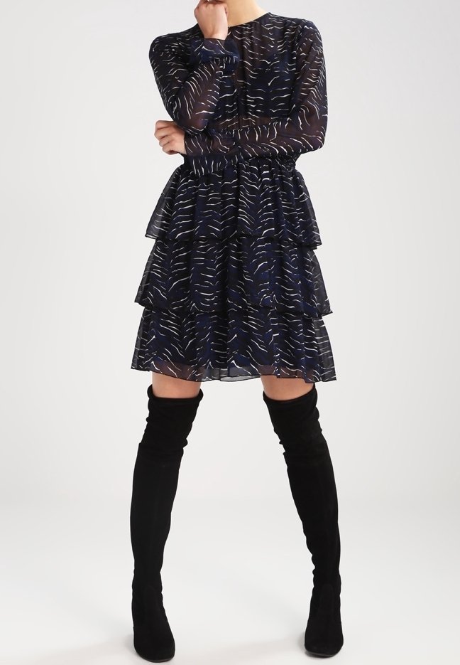 Femme portant une robe bleu marine transparente à manches longues avec une jupe à volants et des rayures blanches abstraites, associée à des bottes noires montant au-dessus du genou.