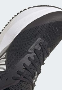 Black athletic shoe featuring a mesh upper, a textured midsole, and a white rubber outsole. Accents include distinct lacing and branding details.