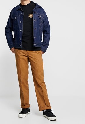 Homme portant une veste en denim bleu foncé, un t-shirt Vans noir, un pantalon couleur camel, et des baskets noires avec des semelles blanches sur un fond uni.