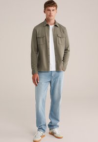 Cămașă verde măsliniu cu nasturi, cu două buzunare pe piept, purtată peste un tricou alb; combinată cu blugi drepți de culoare albastru deschis și adidași gri.