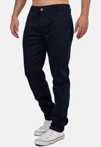 Pantaloni in cotone blu navy con taglio dritto, caratterizzati da dettagli in cucitura rossa e chiusura con bottone, abbinati a sneaker bianche.
