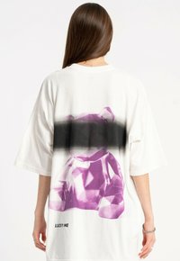 Biała, oversize t-shirt z grafiką fioletowego misia o geometrycznym wzorze i czarnym gradientowym akcentem, z napisem "LUCKY ME" wydrukowanym poniżej.