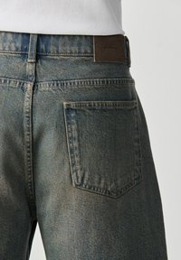 Gros plan d'une personne portant un jean en denim bleu délavé avec une poche arrière et un écusson de marque en cuir marron sur la ceinture.