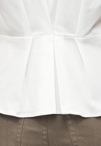 Λεπτομέρεια από λευκό πλισέ ύφασμα πάνω από καφέ υφή υφάσματος, που αναδεικνύει την ραφή και την αντίθεση της δομημένης σχεδίασης.