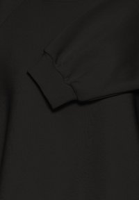 Melna siltinātāja, kam ir mīksta tekstūra, ribotas piedurknes un zirneklim līdzīgas piedurknes. Uzmanība pievērsta bezšuvju dizainam un vienveidīgai krāsai visā apģērbā.