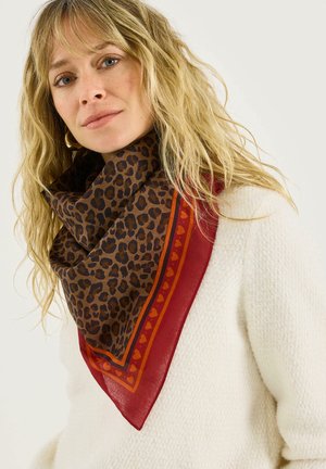 Femme blonde portant un pull blanc texturé et une écharpe à imprimé léopard avec une bordure à motifs de cœurs rouges et orange.