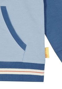Nærbillede af en blå sweatshirtlomme med mørkere blå trim, stribet ribkant og et lille gult stofmærke på sidesømmen.