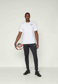 Vīrietis baltā Parīzes Nike T-kreklā, melnās Nike biksēs un melnās kurpēs, turot futbolu, stāv pret vienkāršu gaišu sienu.