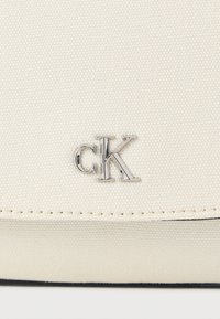 Λευκή υφασμάτινη τσάντα χειρός με λεία υφή, που διαθέτει ασημί λογότυπο "cK" και αντίθετη σκουρόχρωμη γέμιση κατά μήκος του κάτω άκρου.