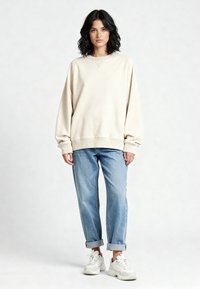 Sweat-shirt beige avec un col rond et des poignets côtelés, associé à un jean large bleu clair et des baskets blanches. Design minimaliste.