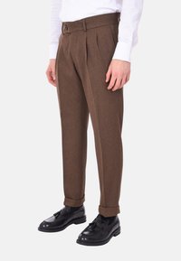 Pantaloni marroni plissettati realizzati in tessuto strutturato, con una vita con bottone e orli risvoltati, abbinati a scarpe nere.