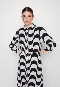 Vestido longo preto e branco com padrão em ondas, com botões na frente, colarinho e mangas três quartos, feito de um tecido leve.