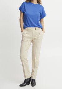 Donna con un top blu a maniche corte, pantaloni beige a vita alta con pieghe frontali e stivali neri con punta affusolata, in piedi con le mani nelle tasche.