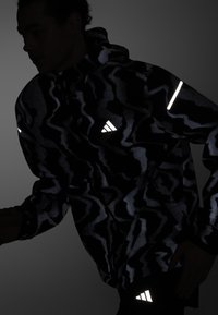 Schwarze reflektierende Jacke mit einem Camouflage-Muster, ausgestattet mit Kapuze, sichtbaren Nähten und Adidas-Logo-Akzenten auf der Brust und dem Ärmel.