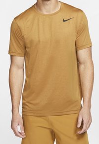 Kurzärmliges, runde Halsausschnitt-T-Shirt aus goldbraunem Stoff mit einem subtilen Glanz, versehen mit einem schwarzen Nike-Logo auf der linken Brust.