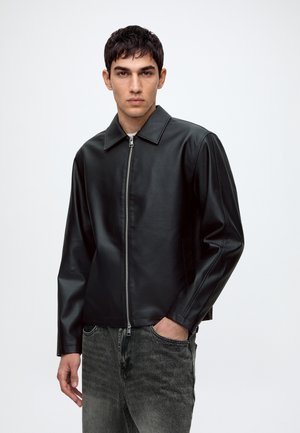 Jeune homme portant une veste en cuir noire zippée avec col et un jean gris délavé, debout avec une main dans la poche devant un fond uni.