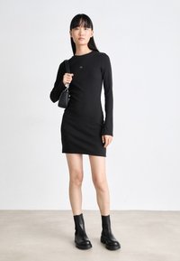Black long-sleeve dress with a fitted silhouette, made of ribbed fabric. Paired with black chunky ankle boots and a small black bag.