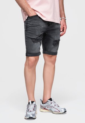 Homme portant un short en jean noir déchiré, un t-shirt rose clair, des baskets argentées et blanches, et un bracelet blanc à perles à son poignet droit.
