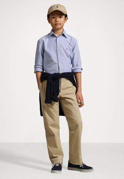 Ανοιχτό μπλε πουκάμισο με γυρισμένα μανίκια, μπεζ παντελόνι, ναυτικό πουλόβερ δεμένο στη μέση, μπεζ καπέλο και σκούρα αθλητικά παπούτσια με άσπρα κορδόνια.