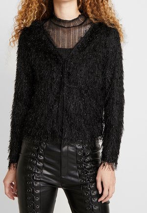 Frau trägt eine schwarze, flauschig strukturierte Strickjacke über einem durchsichtigen, gestreiften schwarzen Oberteil und schwarzen, geschnürten Hosen aus Kunstleder.