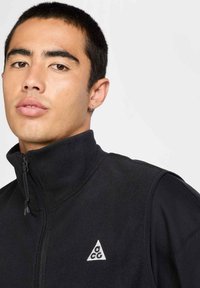 Black fleece vest with high collar and zip, featuring a triangular white logo on the left chest. Smooth texture and fitted design.