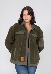 Zielona oliwkowa kurtka polarowa z zapięciem na guziki z przodu, kieszeniami na piersi oraz kieszenią zapinaną na zamek na rękawie. Posiada miękką teksturę i widoczną metkę marki.